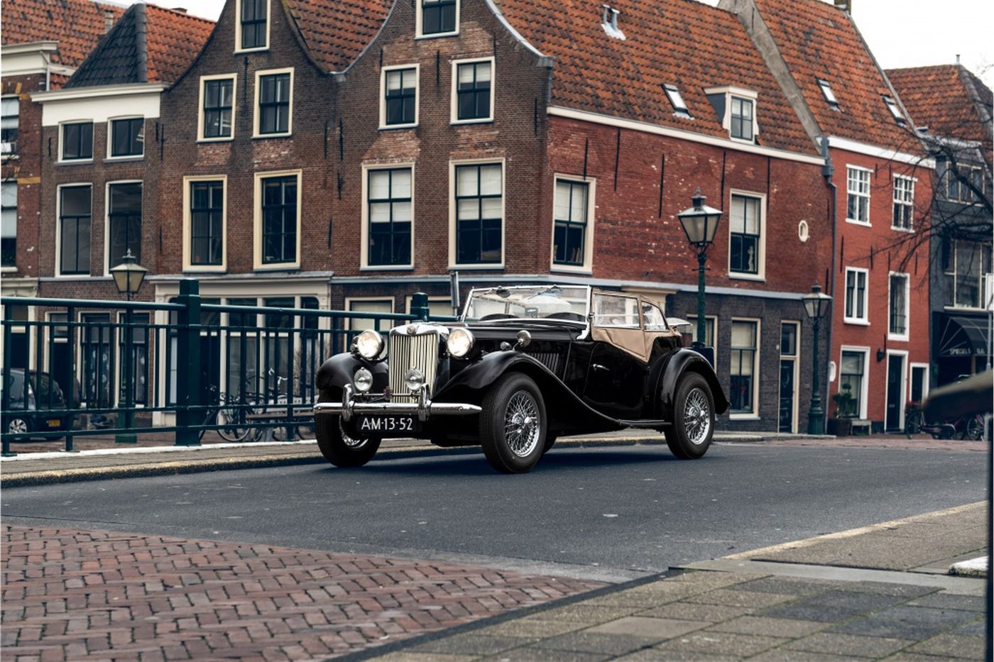 Zwarte MG TD op een brug voor een rij Nederlandse gevels — oldtimer uit nalatenschap